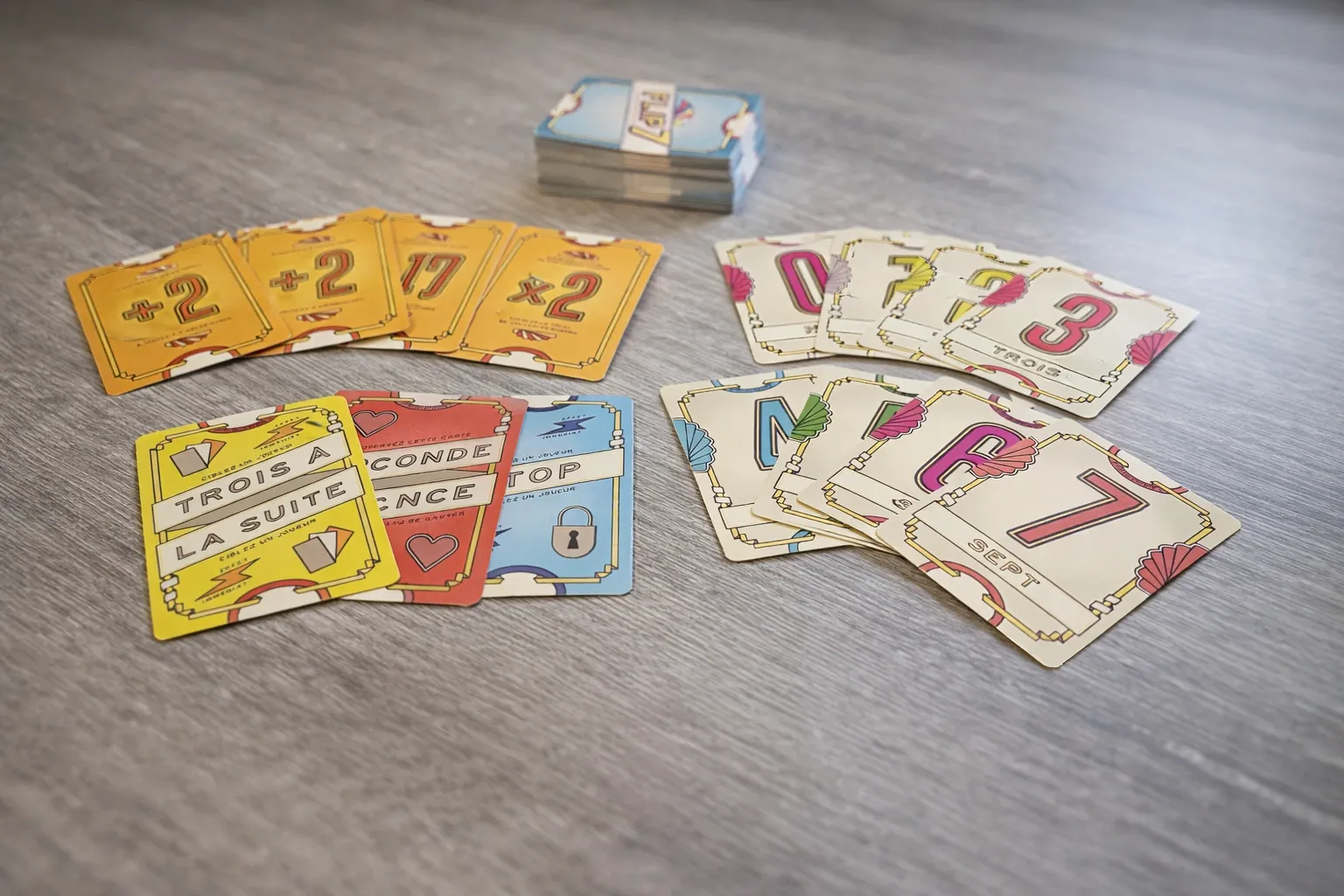 Plusieurs cartes de jeu colorées avec chiffres et symboles sont disposées en éventail sur une table en bois, avec une pile de cartes au fond.
